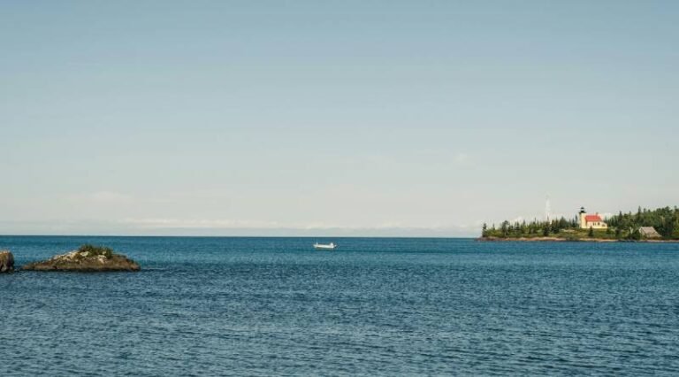 A large body of water with a white boat in the middle and a lighthouse on a crop of land far out.